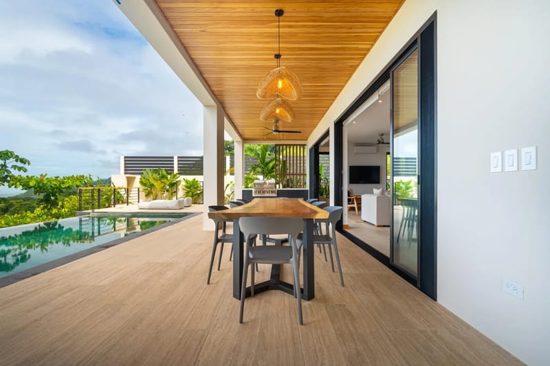 Villa del Cielo · 16. This spacious covered outdoor terrace features a long dining table, an infinity pool, and expansive views of lush greenery and the ocean under a bright sky, providing a seamless transition to the interior living space.