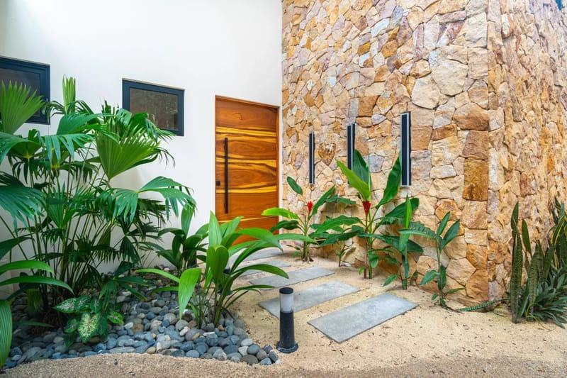 Villa del Cielo · 12. The villa entrance features a prominent wooden door, framed by a natural stone wall, a clean white exterior with windows, and lush tropical plants along a path of concrete stepping stones.