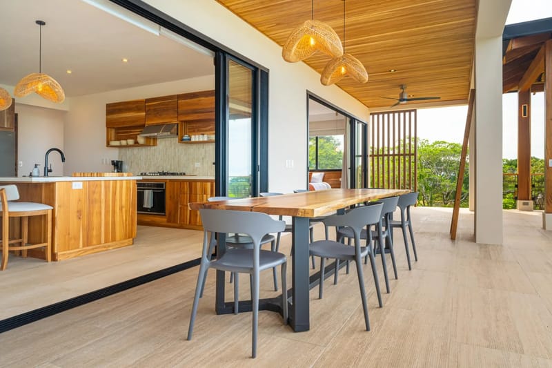 Villa del Cielo · 19. An outdoor terrace features a wooden dining table and grey chairs under a natural wood ceiling, opening to an indoor kitchen via sliding glass doors and offering views of surrounding trees.