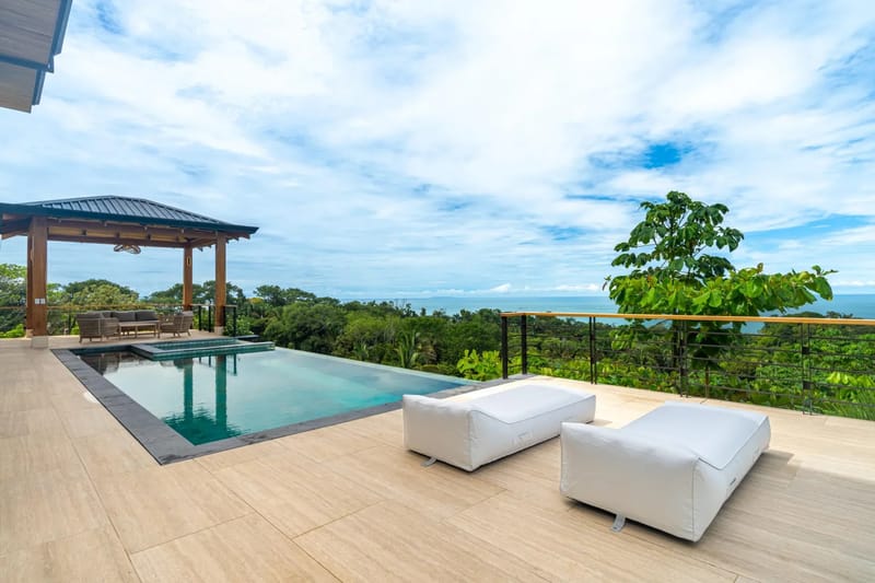 Villa del Cielo · 13. An infinity pool with an integrated jacuzzi is set on a tiled terrace featuring lounge seating and a shaded palapa, all overlooking a vast jungle leading to the ocean.