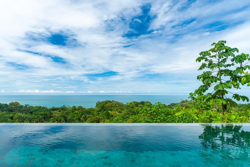 Villa del Cielo · 18. An infinity pool with clear blue water extends towards a panoramic view of the Pacific Ocean and verdant jungle beneath a cloudy sky.