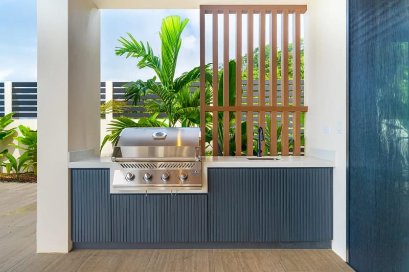 Villa del Cielo · 11. A covered outdoor kitchen features a built-in stainless steel grill and sink, complemented by dark ribbed cabinetry and tropical greenery visible through a wooden slat screen.