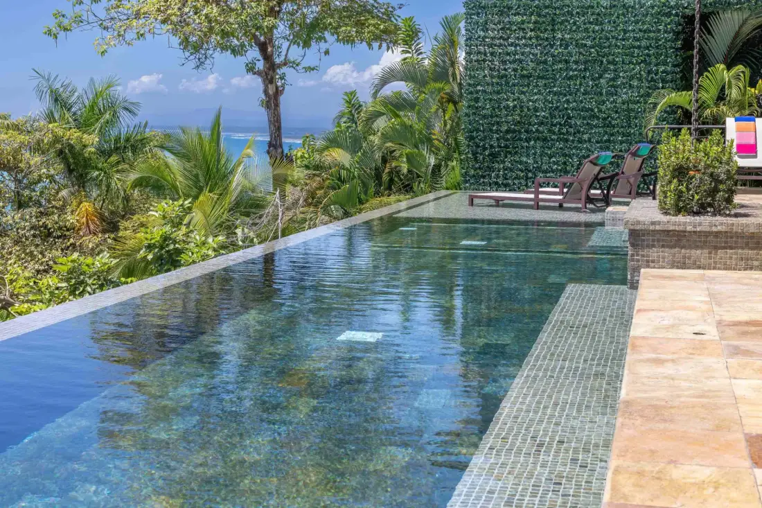 An outdoor swimming pool beside tropical greenery and lounge seating.
