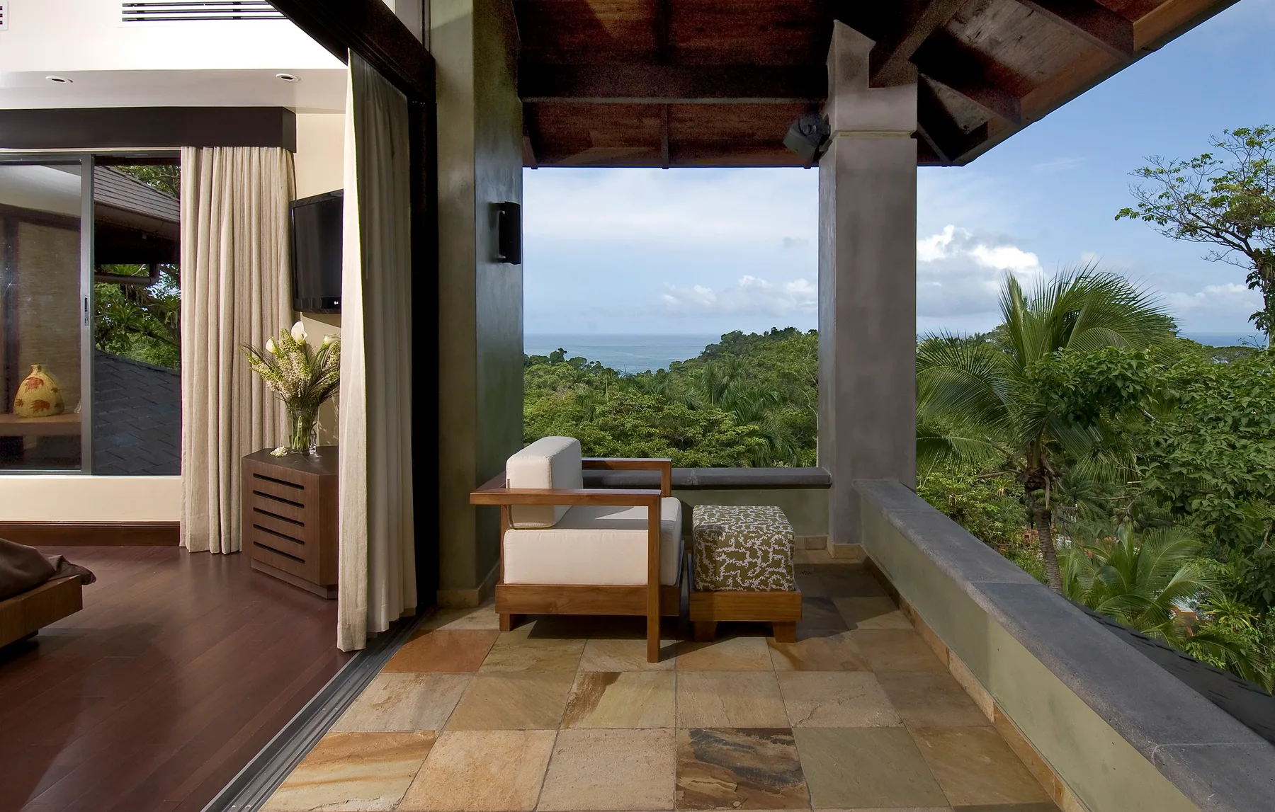 A covered terrace with seating overlooks green treetops and the ocean.