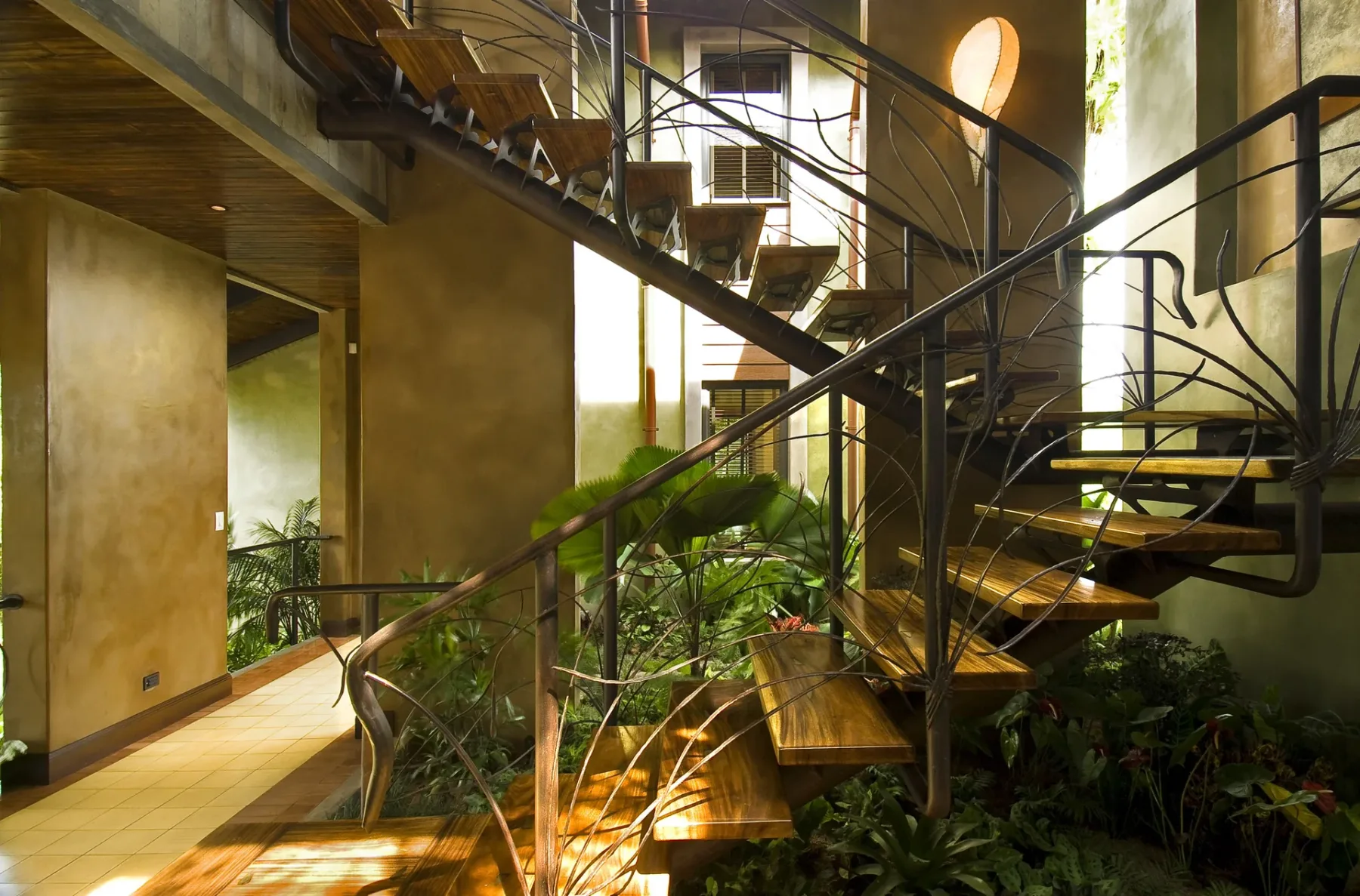 An indoor staircase with open wooden steps and dark metal railings rises above indoor plants.