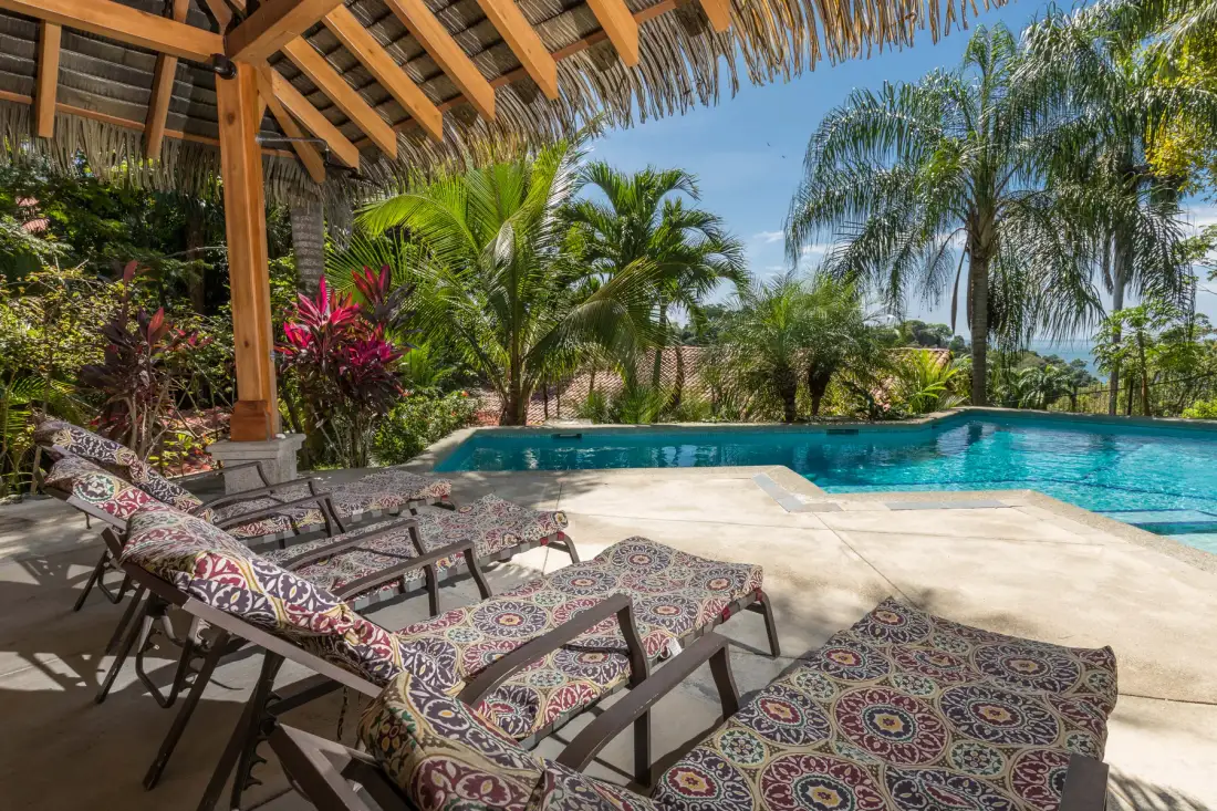 Una piscina junto a un patio sombreado con asientos reclinables y plantas tropicales.