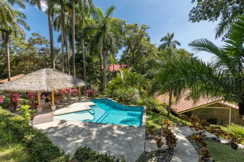 Casa Colibrí · 25. El espacio exterior incluye una piscina y una palapa con techo de palma, enmarcadas por altas palmeras y vegetación tropical bajo un cielo luminoso.