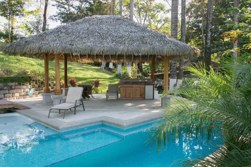 Casa Colibrí · 26. Una gran piscina revestida de azulejos azules y una terraza se encuentran junto a un pabellón con techo de palma y una barra integrada, entre vegetación tropical y una ladera verde.