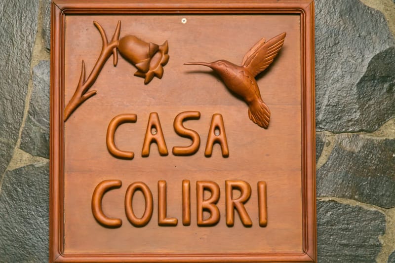 Casa Colibrí · 28. Un letrero de madera hecho a mano de Casa Colibri, con un colibrí y una flor tallados, está colocado sobre una pared de piedra con textura.