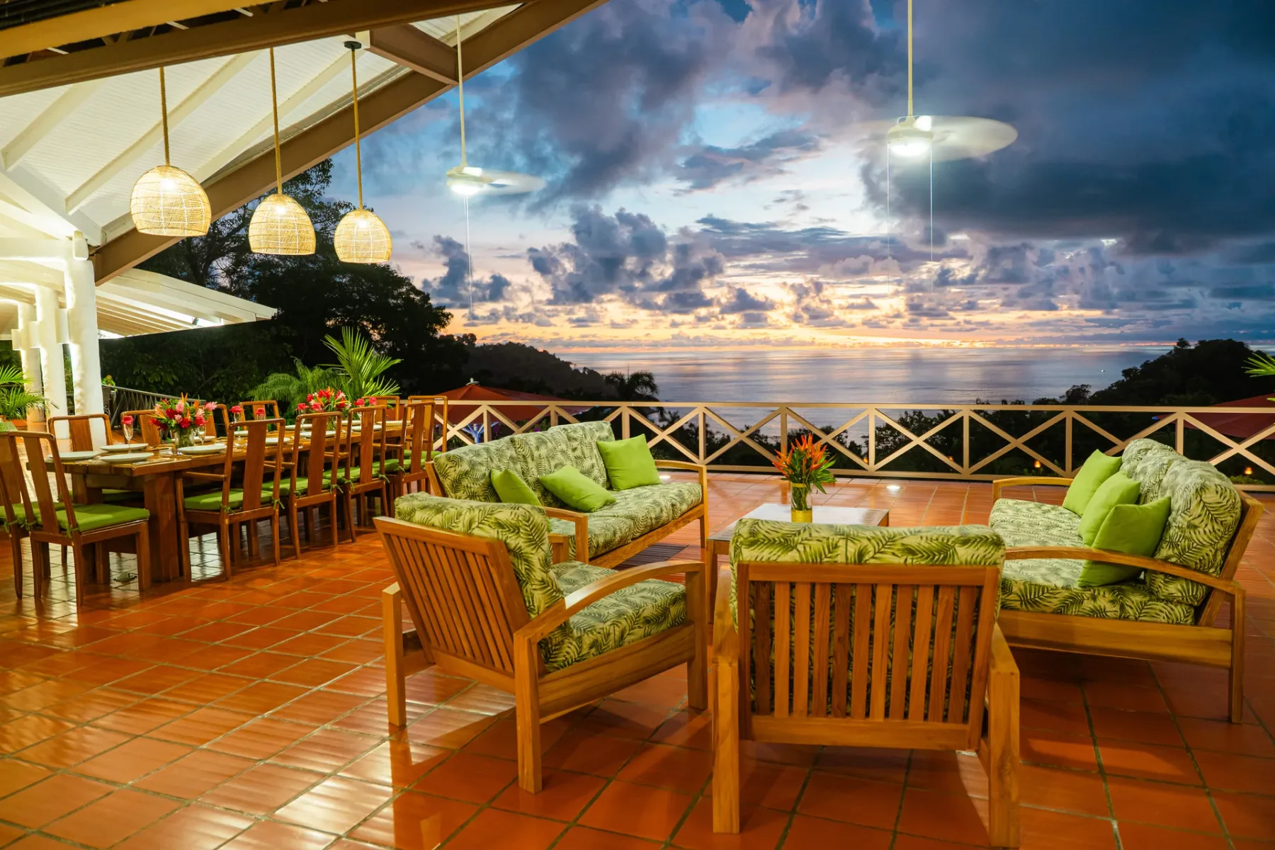 Una terraza al aire libre tiene asientos de madera y una mesa de comedor frente al mar al atardecer.