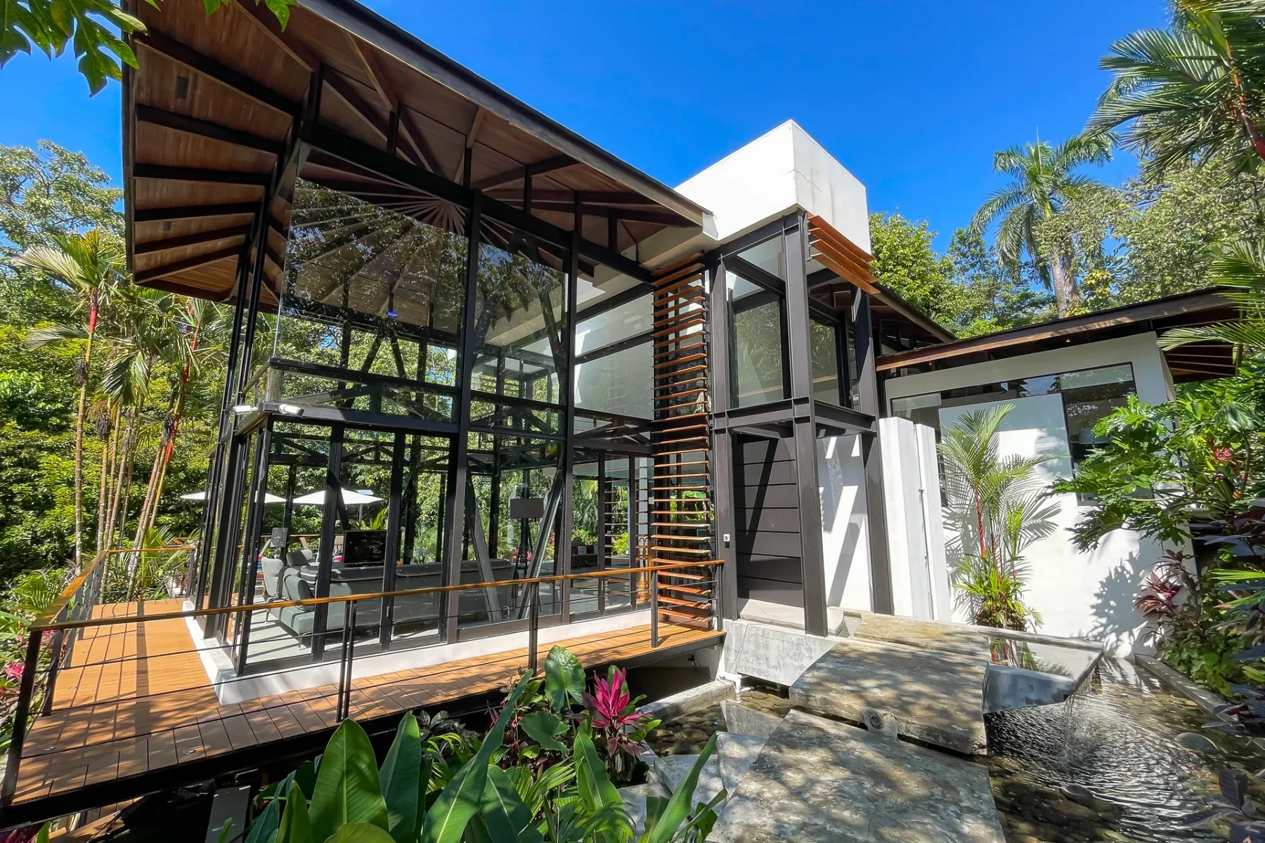 Una villa moderna con fachada de vidrio está rodeada de vegetación tropical bajo un cielo azul despejado.