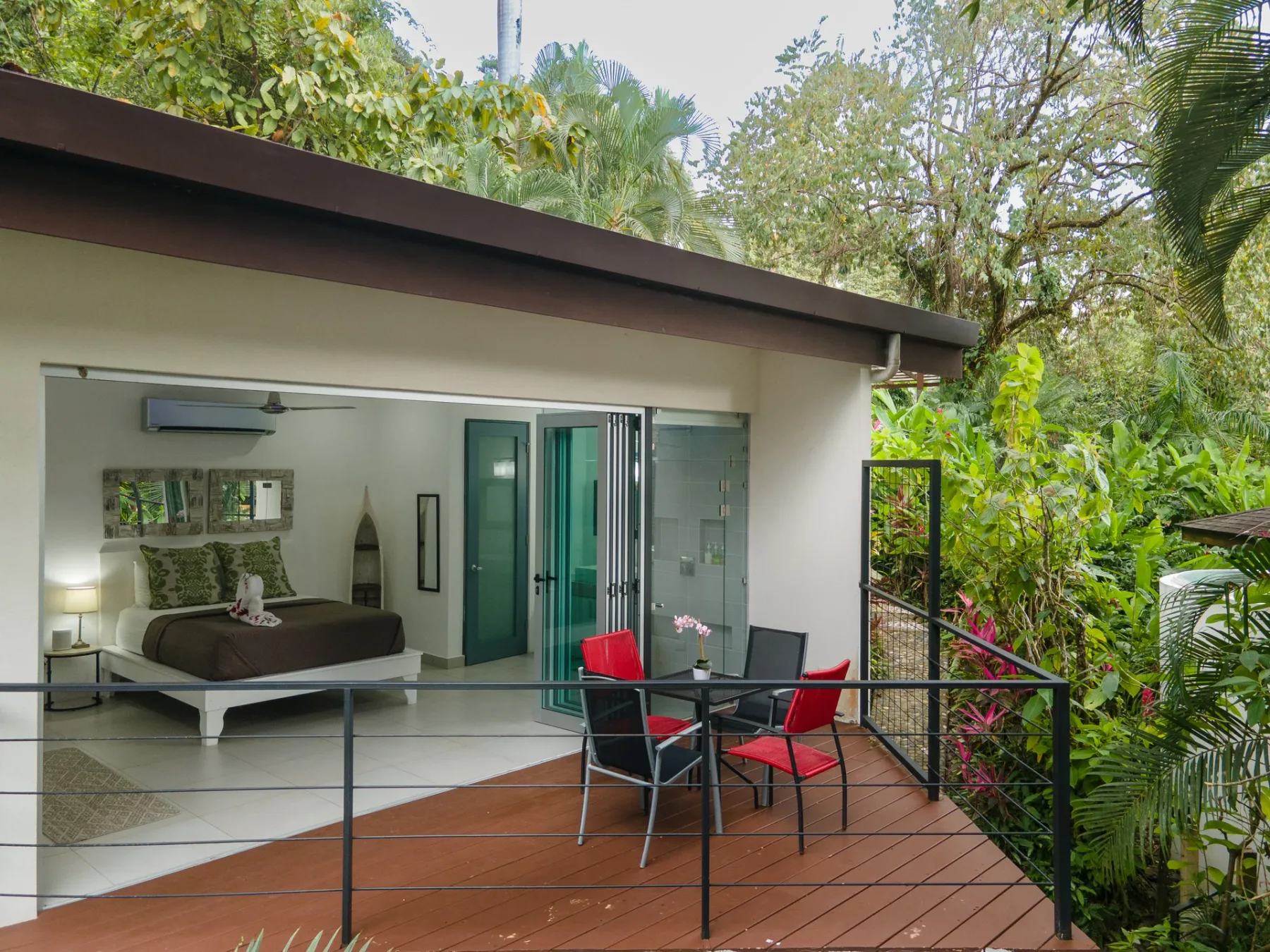 Un dormitorio se abre a una terraza de madera con asientos rojos y vegetación alrededor.