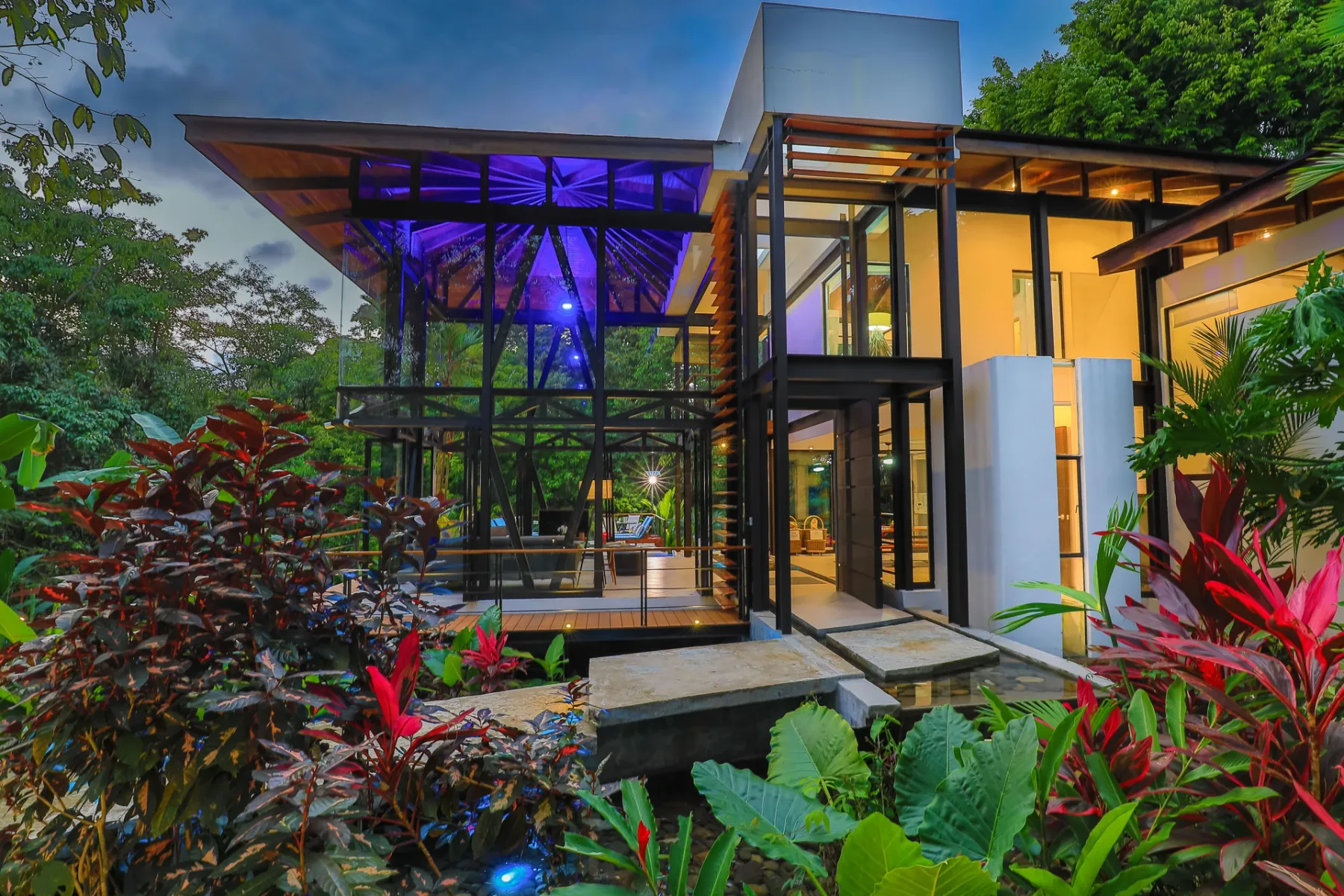 Exterior de una casa moderna con paredes de vidrio y vegetación tropical.