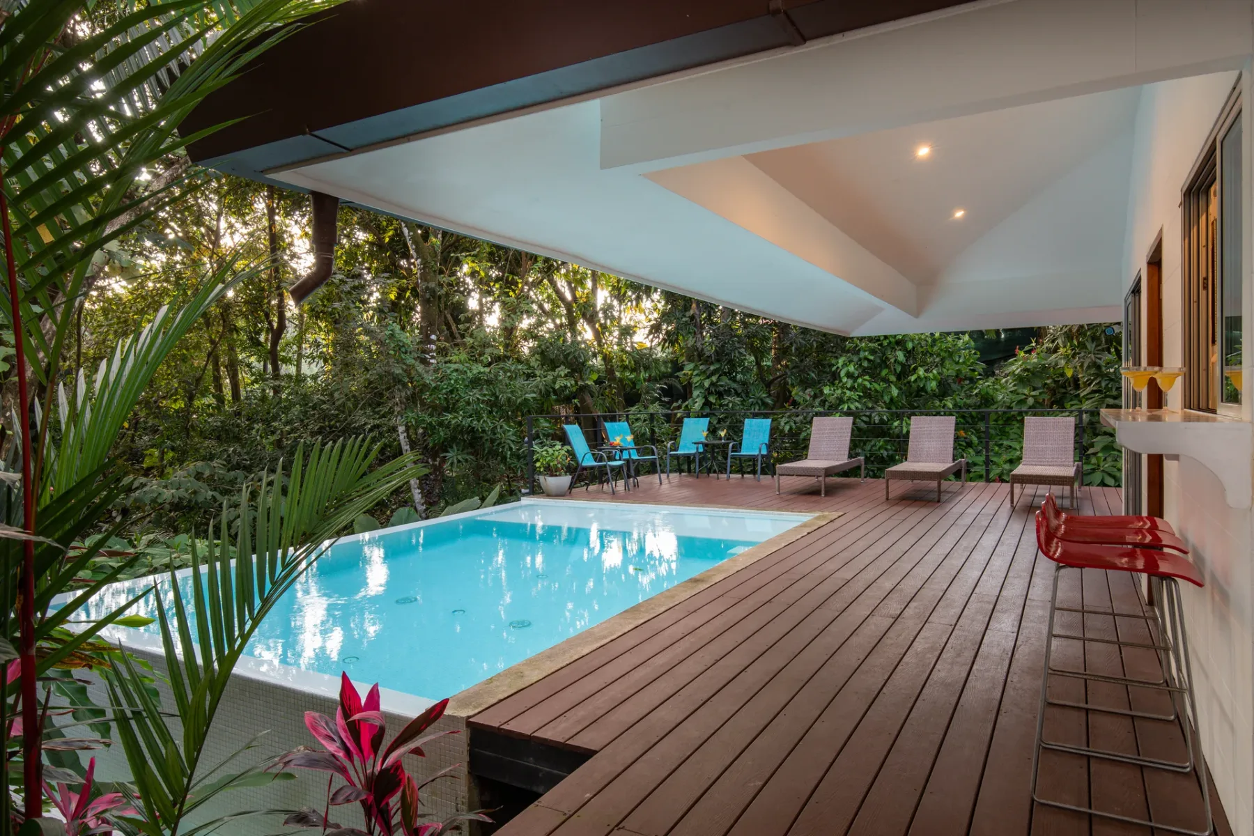 Una terraza cubierta junto a la piscina, con asientos y vegetación alrededor.