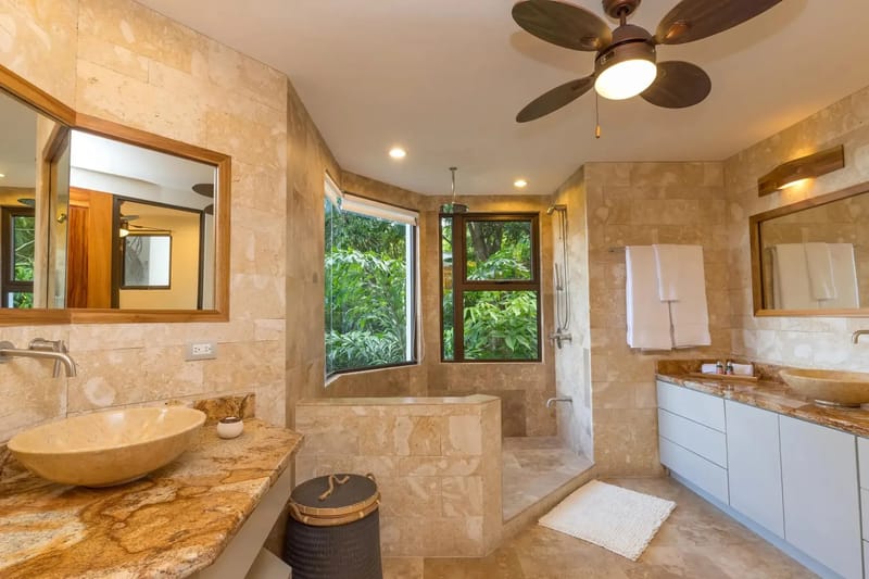 Casa Anjali Luxury Villa · 23. A stone-tiled bathroom features dual granite vanities with vessel sinks, an integrated shower and soaking area, and large windows inviting in natural light and tropical rainforest views.