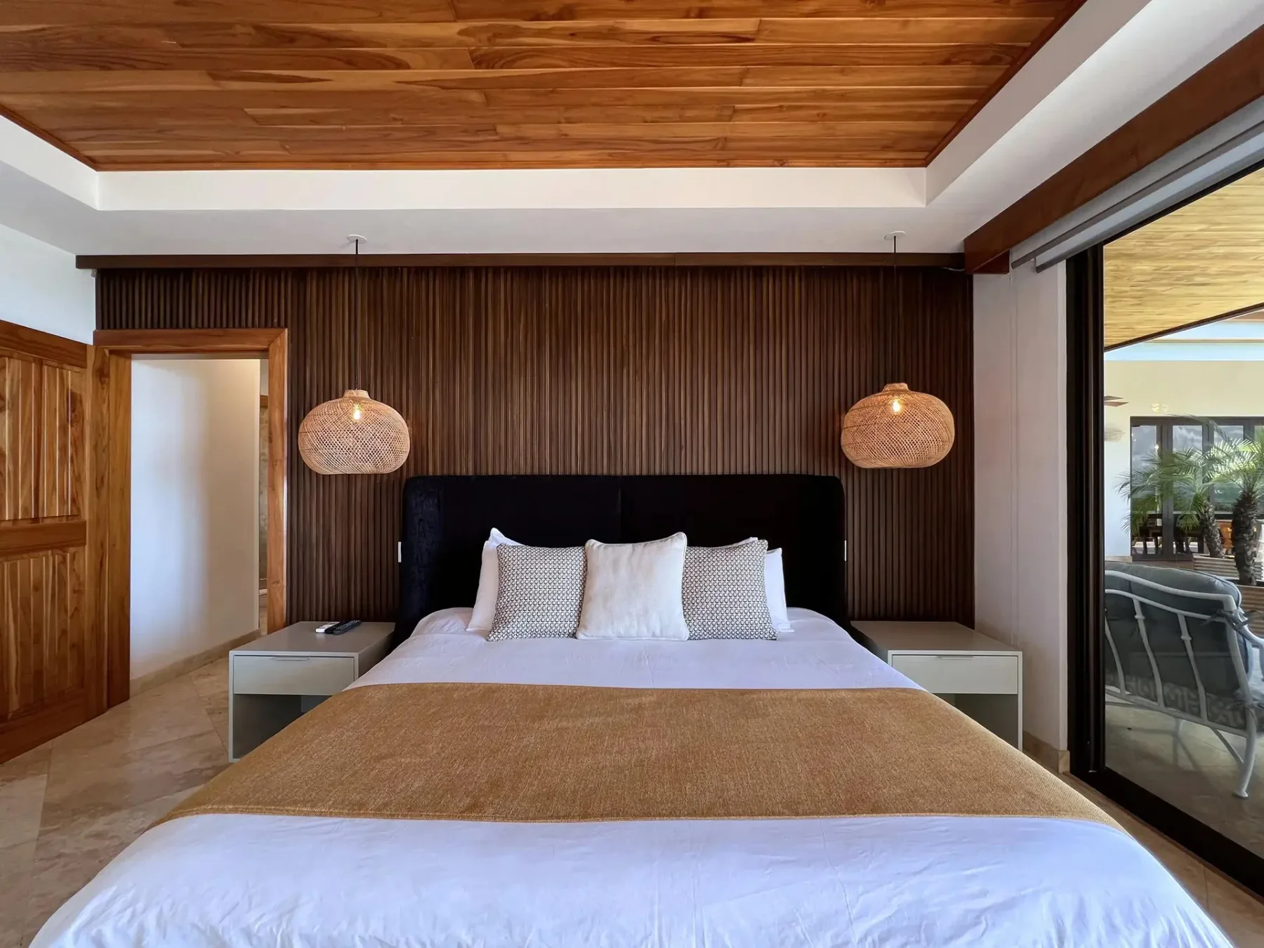 A bedroom with a bed beneath a wood-paneled ceiling, beside large glass doors.
