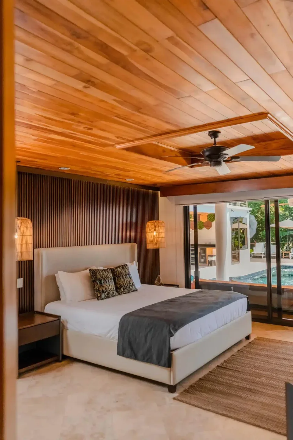 Casa Anjali Luxury Villa · 61. This bedroom features a wood-paneled ceiling with a fan, an accent wall with vertical wood slats, and large glass doors offering natural light and a view of the poolside and tropical surroundings.