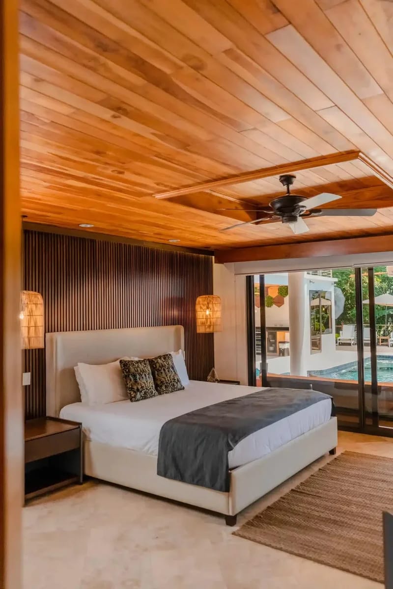 Casa Anjali Luxury Villa · 61. This bedroom features a wood-paneled ceiling with a fan, an accent wall with vertical wood slats, and large glass doors offering natural light and a view of the poolside and tropical surroundings.