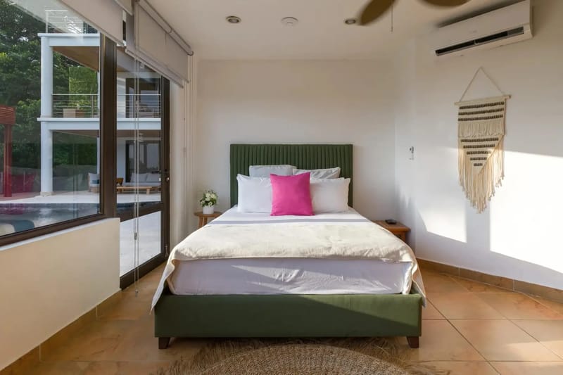 Casa Anjali Luxury Villa · 72. This bright bedroom features a king bed, tiled floors, and floor-to-ceiling glass doors providing direct access to the pool terrace and views of the multi-level villa and tropical foliage.