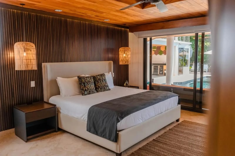 Casa Anjali Luxury Villa · 60. This bedroom offers a king bed under a wooden ceiling, a slatted headboard wall, and sliding glass doors that frame a view of the adjacent pool and tropical landscape.