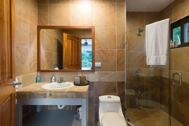 Casa Anjali Luxury Villa · 69. A bathroom with warm-toned tiled walls features a stone vanity, a glass-enclosed shower with a rainforest view, and a wooden door opening to an adjacent space with natural light.