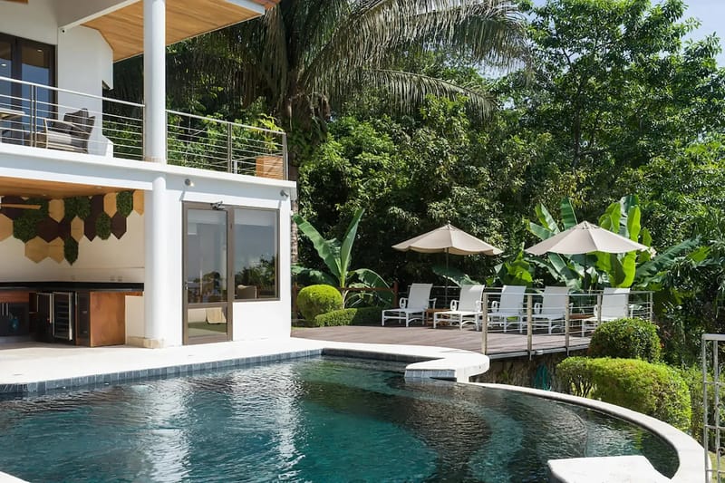 Casa Anjali Luxury Villa · 19. A multi-level villa with an outdoor covered kitchen area overlooks a dark-tiled swimming pool and sun deck with lounge chairs, all surrounded by vibrant tropical rainforest.