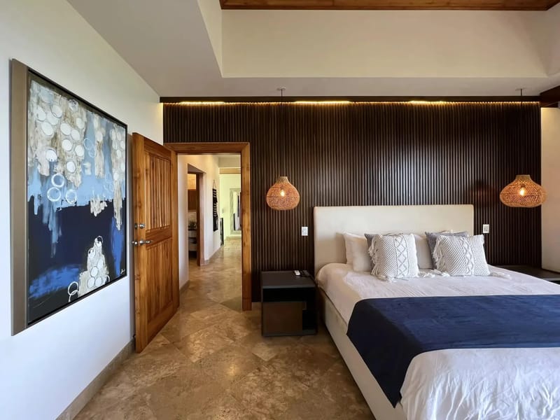 Casa Anjali Luxury Villa · 24. This bedroom features a king bed with an upholstered headboard against a dark wood slatted wall, illuminated by cove lighting and woven pendant lamps, set on light-toned tiled flooring, with an open doorway leading to a hallway.
