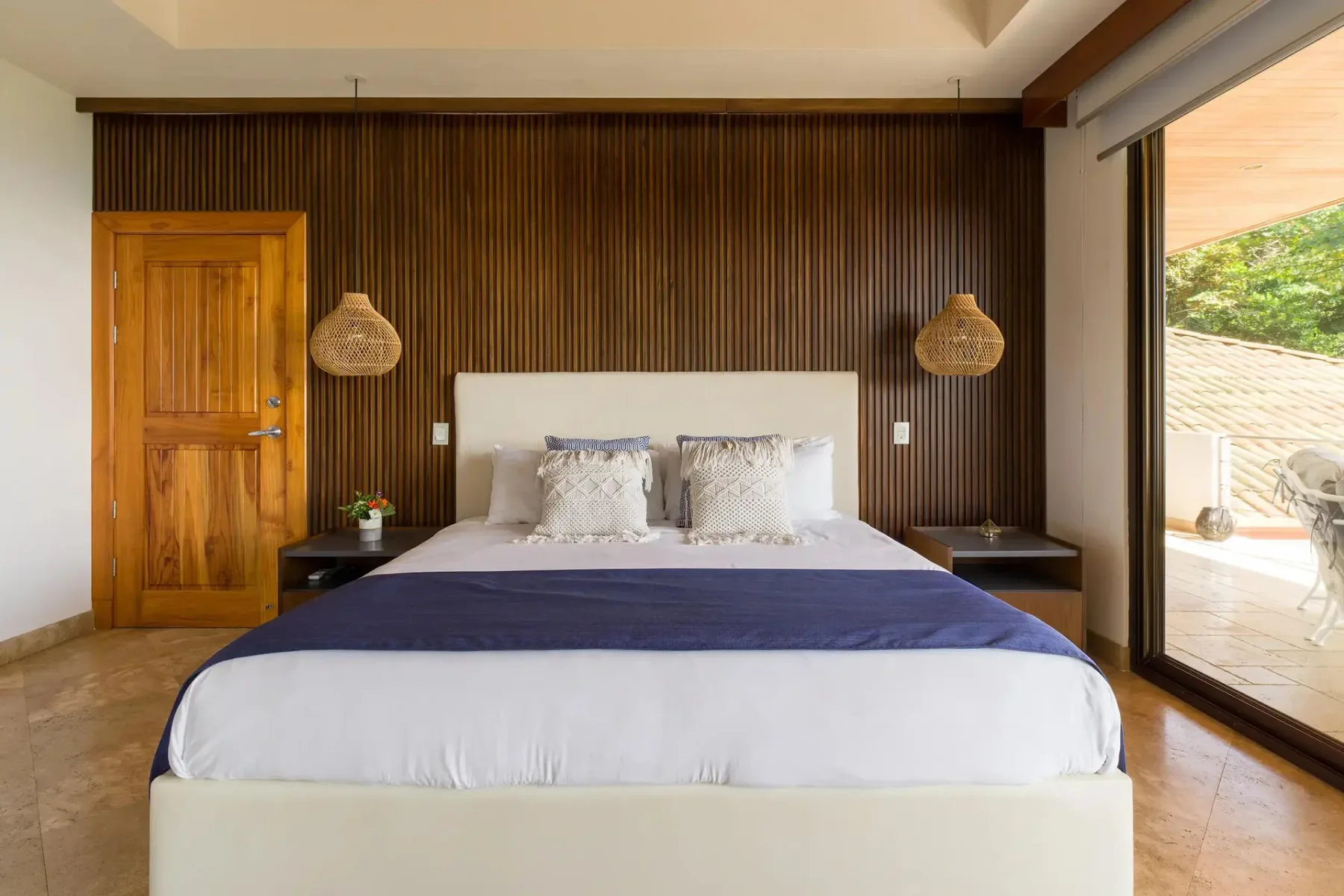 A bedroom with a large bed against a dark slatted wall and a glass opening to the patio.