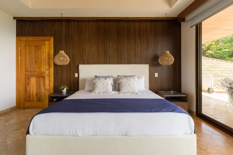 Casa Anjali Luxury Villa · 26. A king bedroom with a slatted wood accent wall and natural woven pendant lights opens to a private terrace through floor-to-ceiling glass doors, offering rainforest views and abundant natural light.