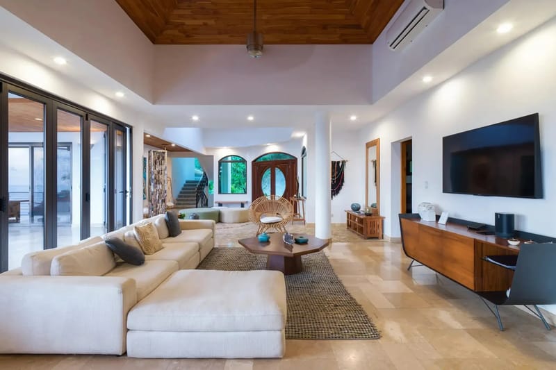 Casa Anjali Luxury Villa · 16. The open-plan living room features a vaulted teak ceiling, light-colored tiled floors, and nine-foot folding glass walls offering views of the ocean and rainforest.