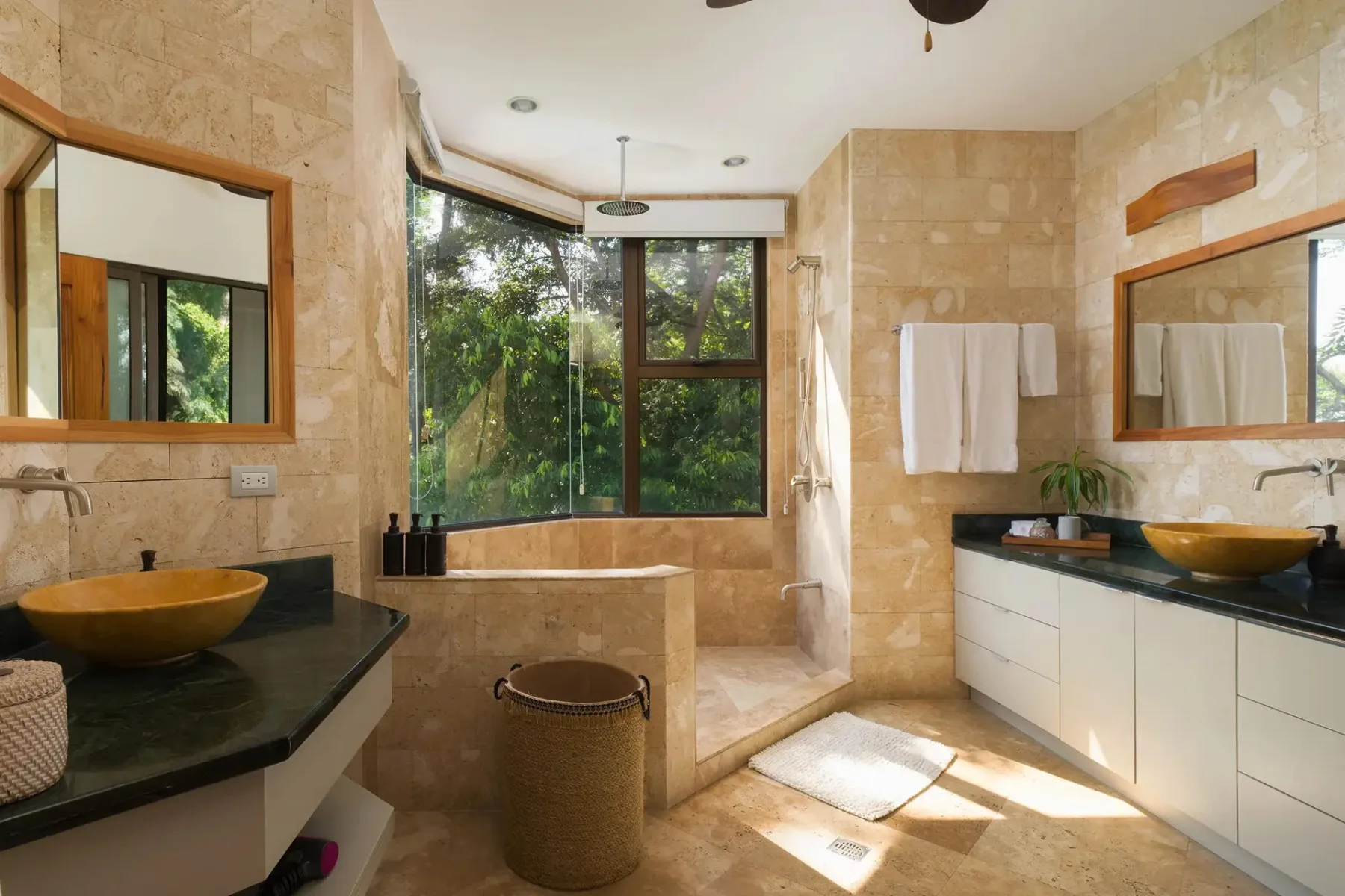 A bright bathroom has stone walls, counter seating, and a large window overlooking greenery.