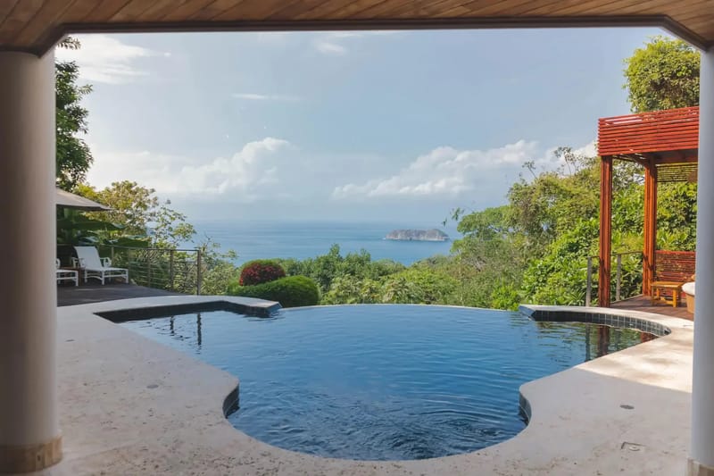 Casa Anjali Luxury Villa · 12. An infinity pool offers an expansive ocean view with a distant island, framed by lush tropical foliage and an adjacent shaded deck.