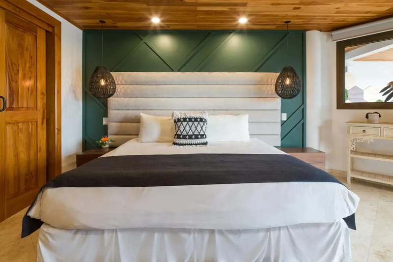 Casa Anjali Luxury Villa · 68. This bedroom features a king bed with an upholstered headboard against a green paneled wall, a wood-paneled ceiling, and a window providing natural light and a glimpse of the tropical surroundings.