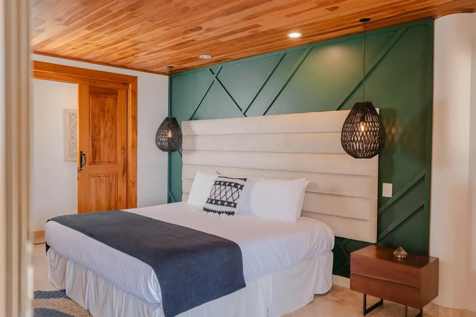 Casa Anjali Luxury Villa · 66. This bedroom features a king bed with a cream upholstered headboard set against a dark green geometric accent wall, beneath a natural wood plank ceiling and flanked by woven pendant lights.