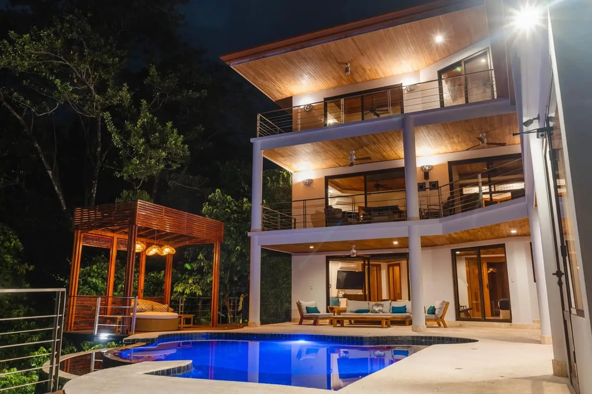 Casa Anjali Luxury Villa · 8. The three-level villa with its wood-paneled terraces and glass doors is lit at night, featuring an illuminated infinity pool and outdoor seating areas amidst the rainforest.
