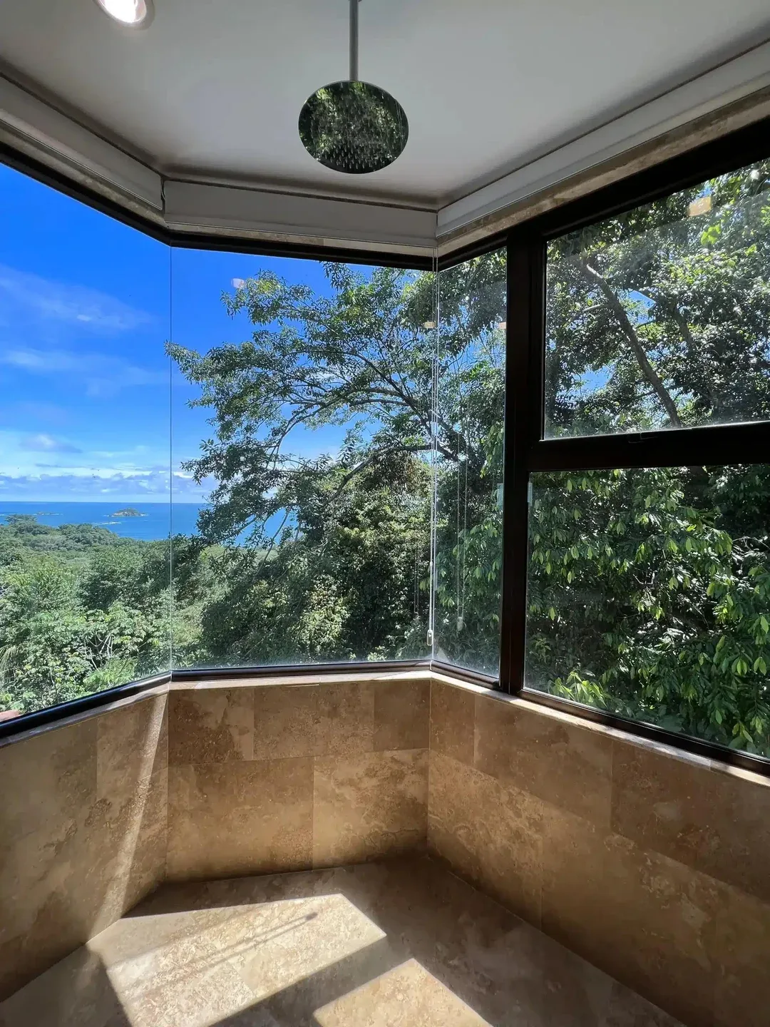 A corner shower area with tan tiled walls and expansive windows overlooking greenery and the ocean.