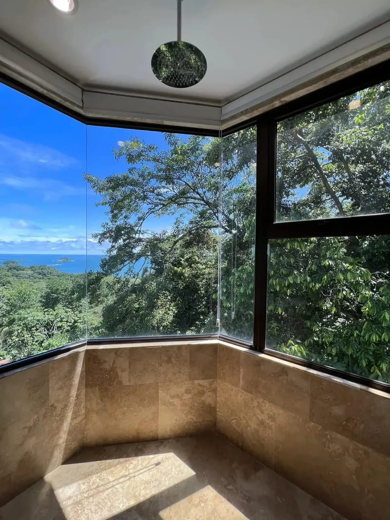 Casa Anjali Luxury Villa · 48. This spacious corner shower features natural stone tile walls and floor, complemented by floor-to-ceiling glass panels offering panoramic views of the ocean and surrounding rainforest canopy.