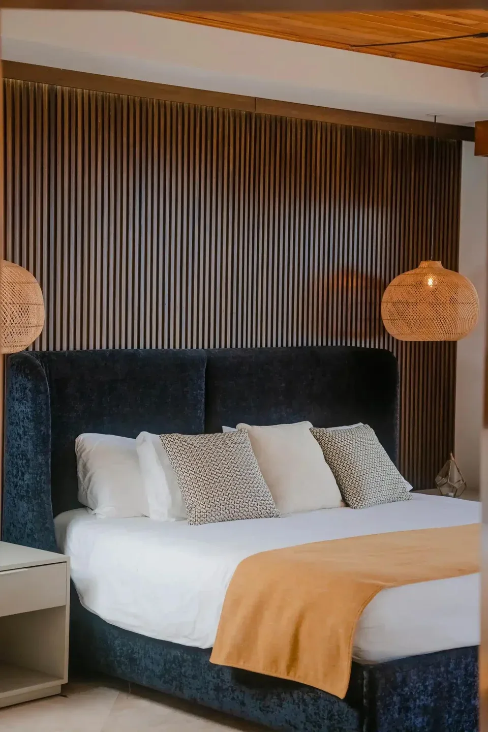 A bedroom with a dark upholstered bed, pale bedding, and woven pendant lights.
