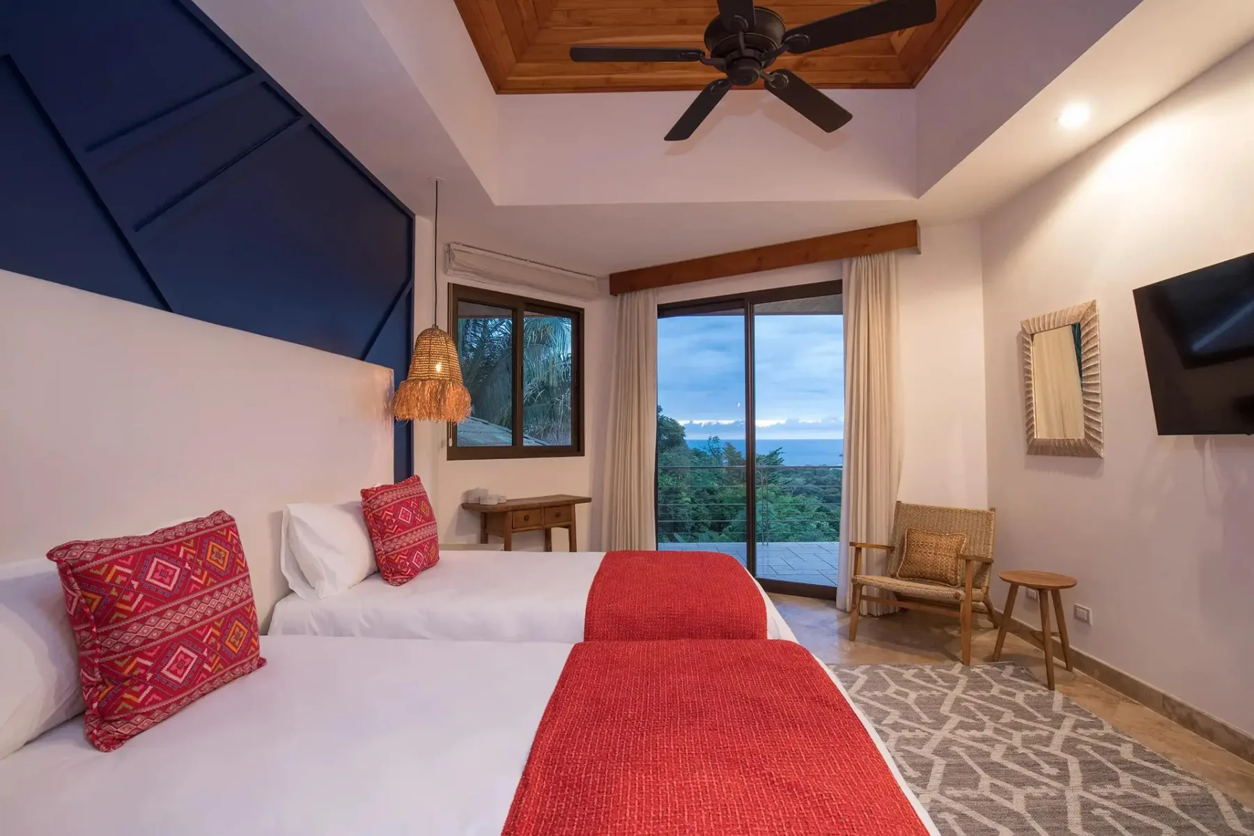 A bedroom with two beds, red bed covers, and glass doors overlooking greenery and water.