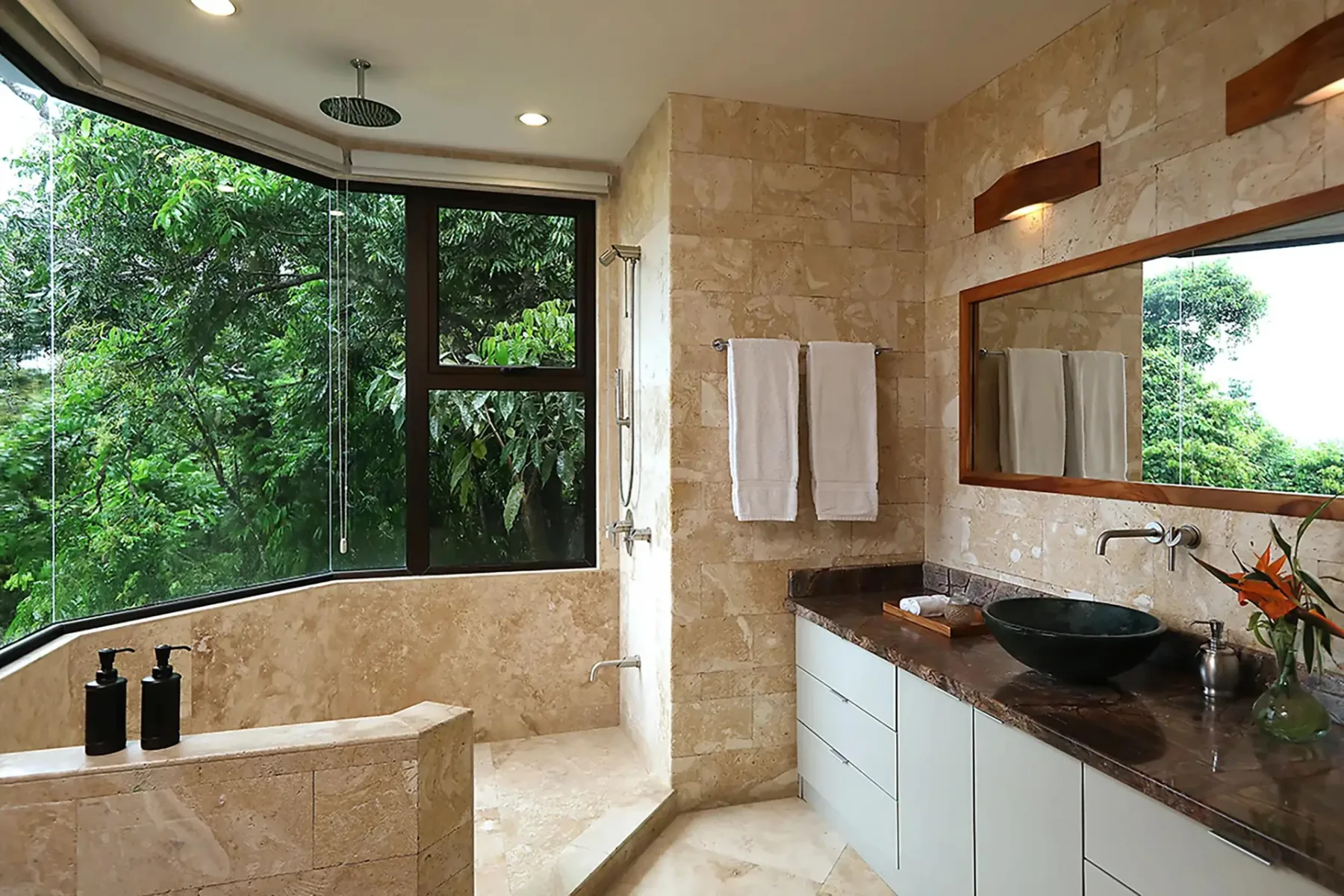 A bathroom with a glass shower, a vanity, and large windows overlooking greenery.