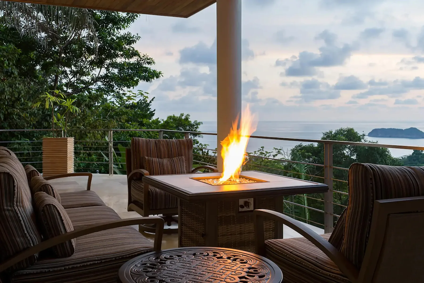 A covered terrace has cushioned seating around a lit fire pit, overlooking the water.