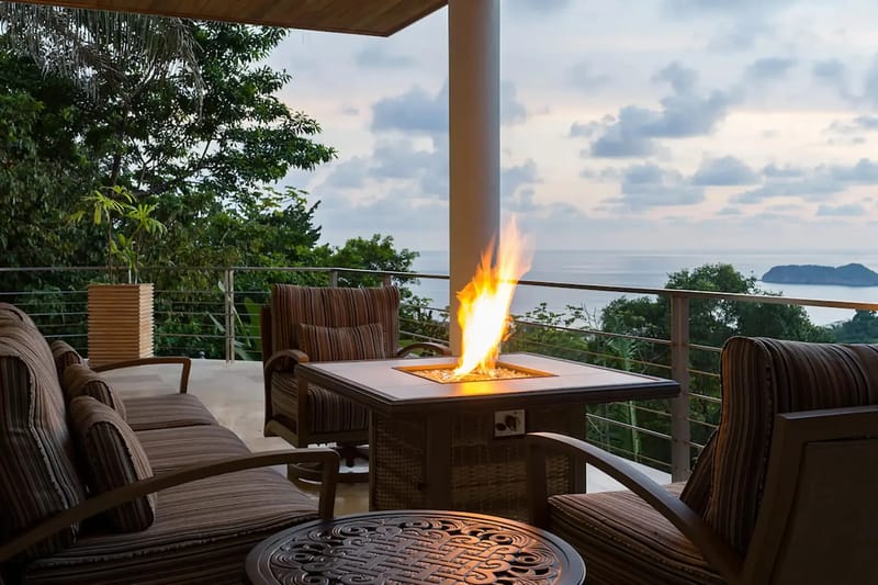 Casa Anjali Luxury Villa · 18. A covered terrace features comfortable cushioned seating and a central lit fire pit, offering broad ocean and rainforest views under a twilight sky.