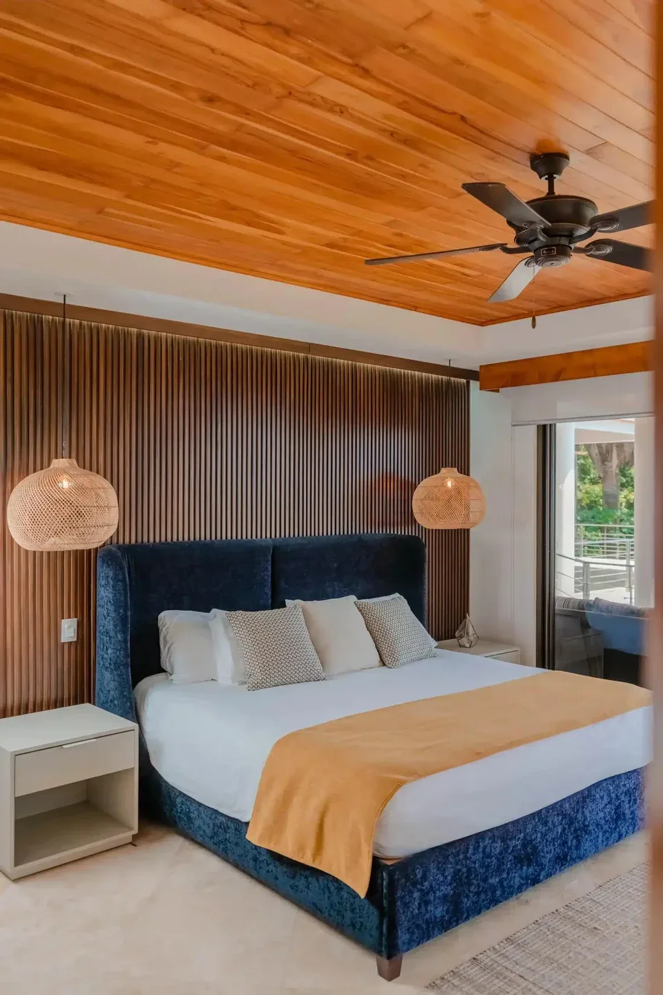 A bedroom with a blue upholstered bed, a slatted wood wall, and a ceiling fan.