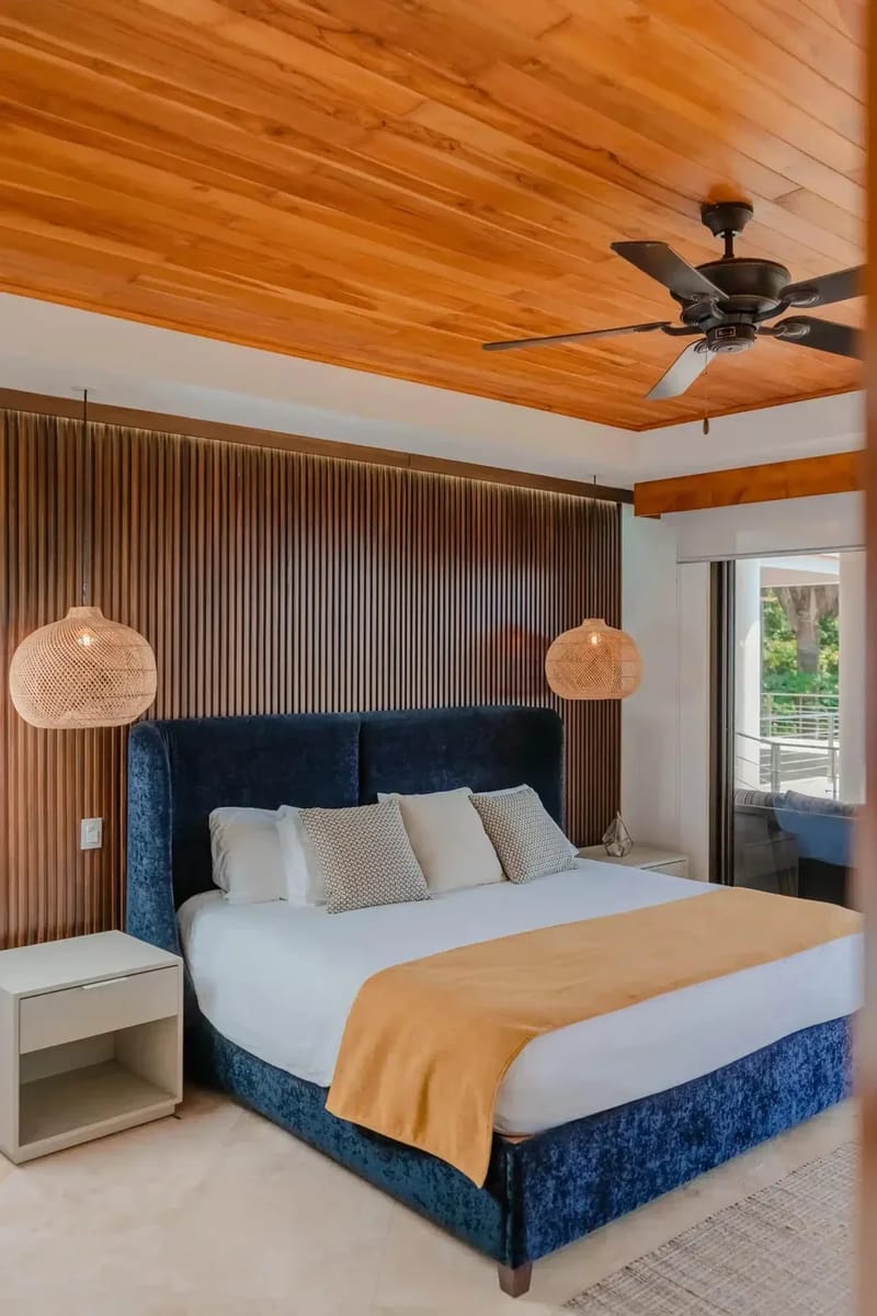 Casa Anjali Luxury Villa · 41. A king bedroom features a blue velvet headboard against a slatted wood accent wall, a warm wood plank ceiling with a ceiling fan, and floor-to-ceiling glass doors that open to a rainforest balcony.