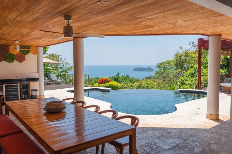 Casa Anjali Luxury Villa · 11. A covered outdoor dining and drink station area features a wooden slatted ceiling and ceiling fans, opening directly to an infinity pool with expansive ocean views and tropical rainforest.