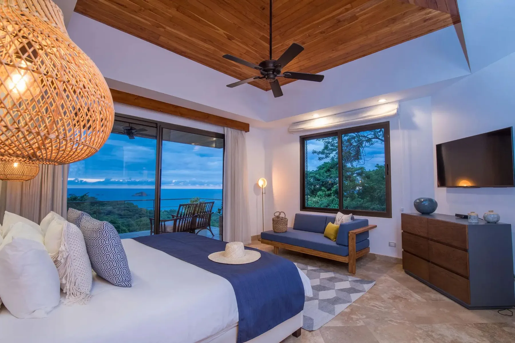 A bedroom with a bed, a wooden ceiling, and glass doors opening onto a balcony with an ocean view.
