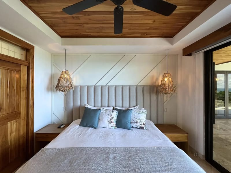 Casa Anjali Luxury Villa · 57. This light-filled bedroom features a wood-paneled ceiling, a king-size bed with an upholstered headboard, and floor-to-ceiling glass doors opening onto a private terrace with ocean and sky views.
