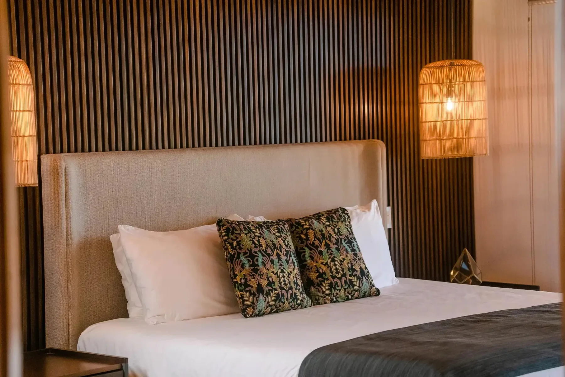 A bedroom with a bed, upholstered headboard, and warm pendant lighting against a slatted wall.