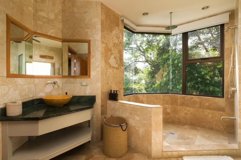 Casa Anjali Luxury Villa · 64. This bathroom, clad in natural stone, features a vanity with a vessel sink and an open shower area, drawing abundant natural light and immersive rainforest views through its expansive corner windows.