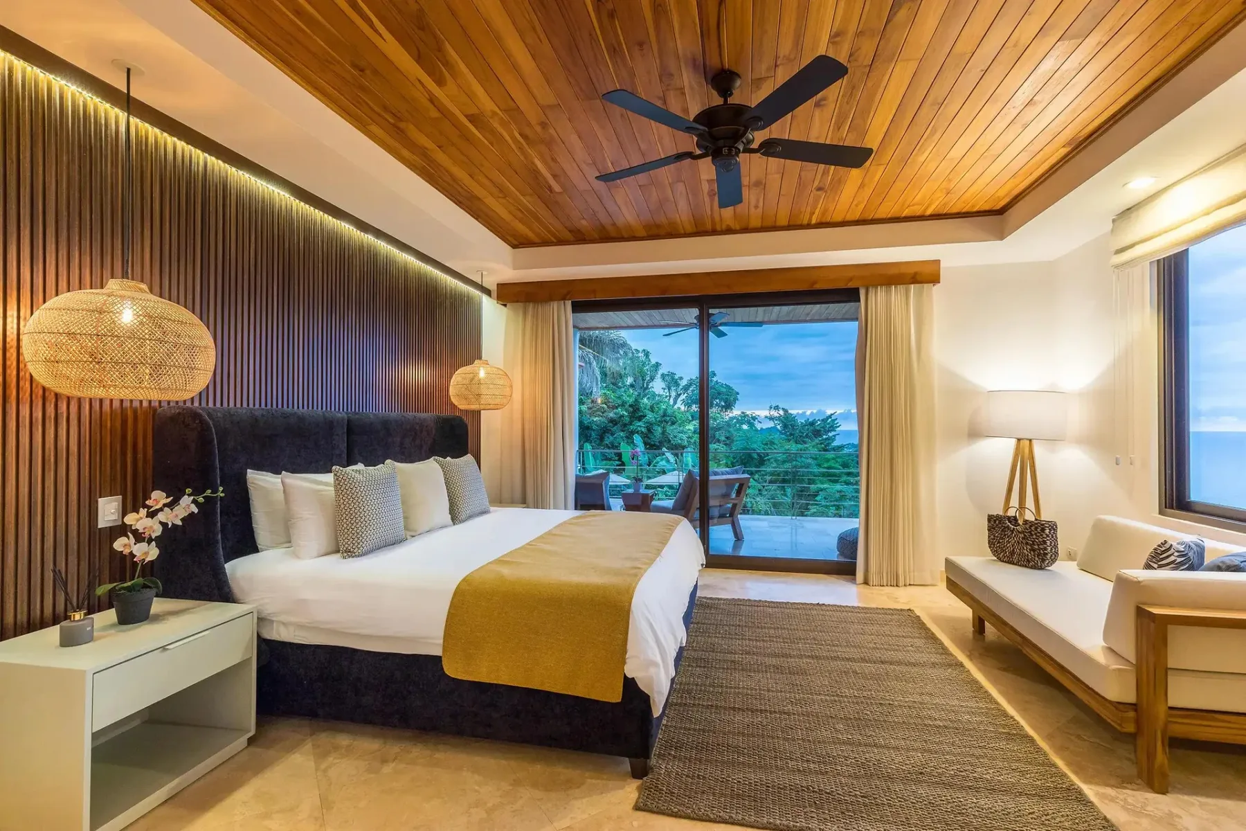 A bedroom with a bed, a wooden ceiling, and glass doors overlooking greenery.